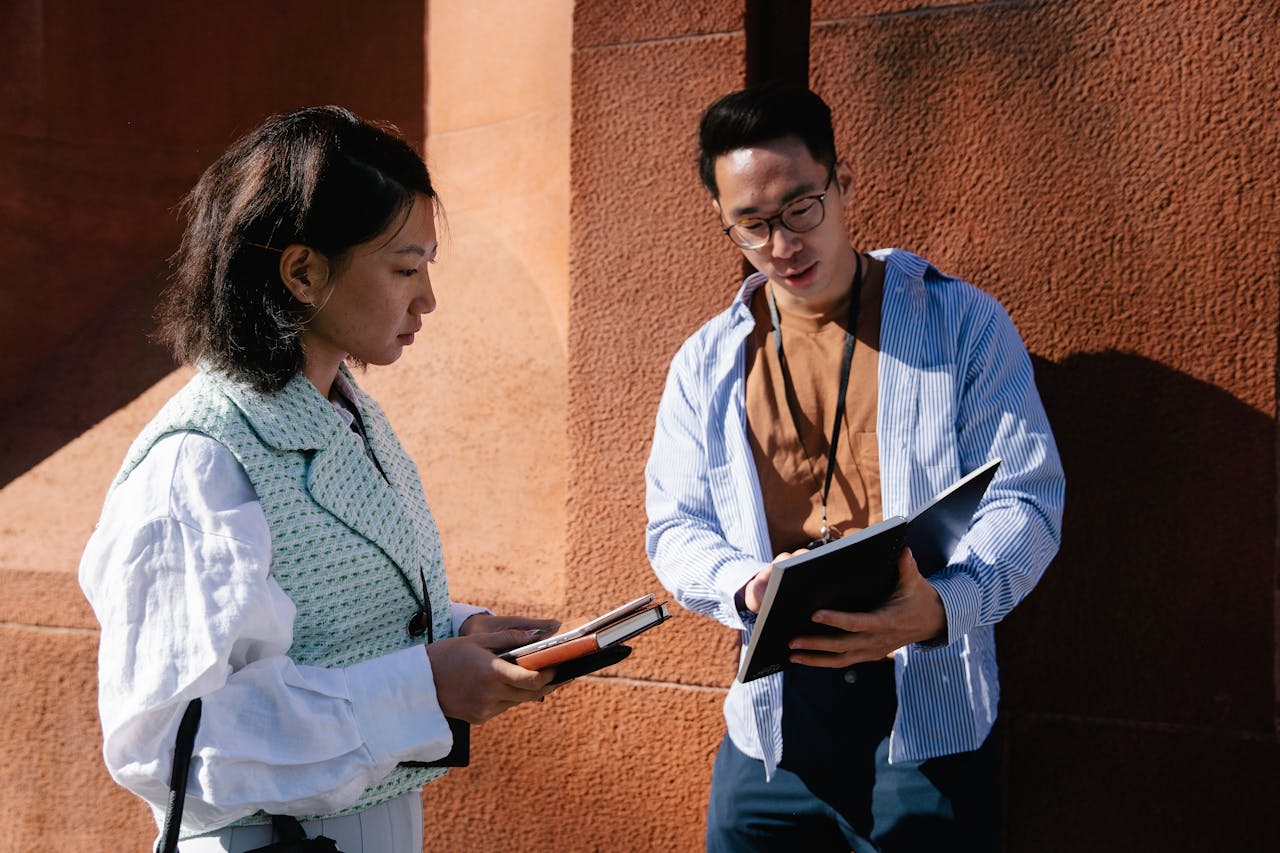 Um homem e uma mulher de aparência oriental. A mulher segura um livro e um tablet nas mãos enquanto o homem, que possuí um crachá no pescoço, apresenta um livro aberto mostrando para a mulher como se estivesse realizando uma entrevista de opinião.